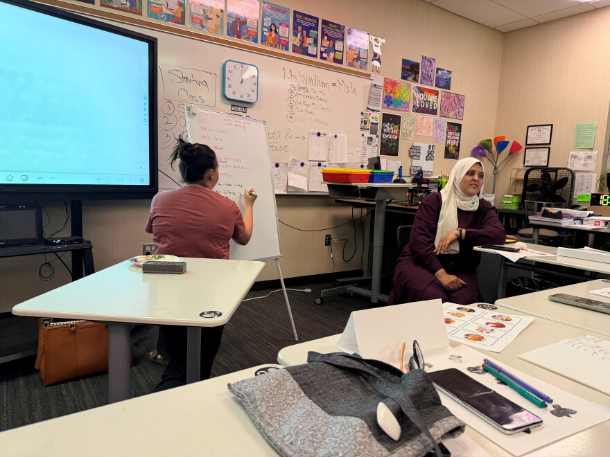 A woman on the left is writing on a whiteboard. To her right, a woman wearing a hijab talks to her class.