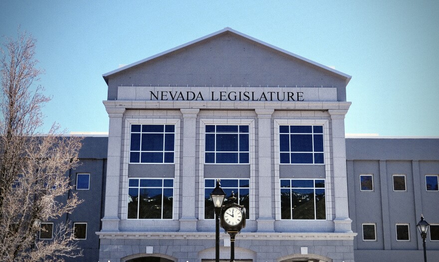 The front facade of the Nevada Legislature building in Carson City, Nevada, photographed on a clear, sunny day. A traditional black clock stands in the foreground, flanked by vintage-style lamp posts.