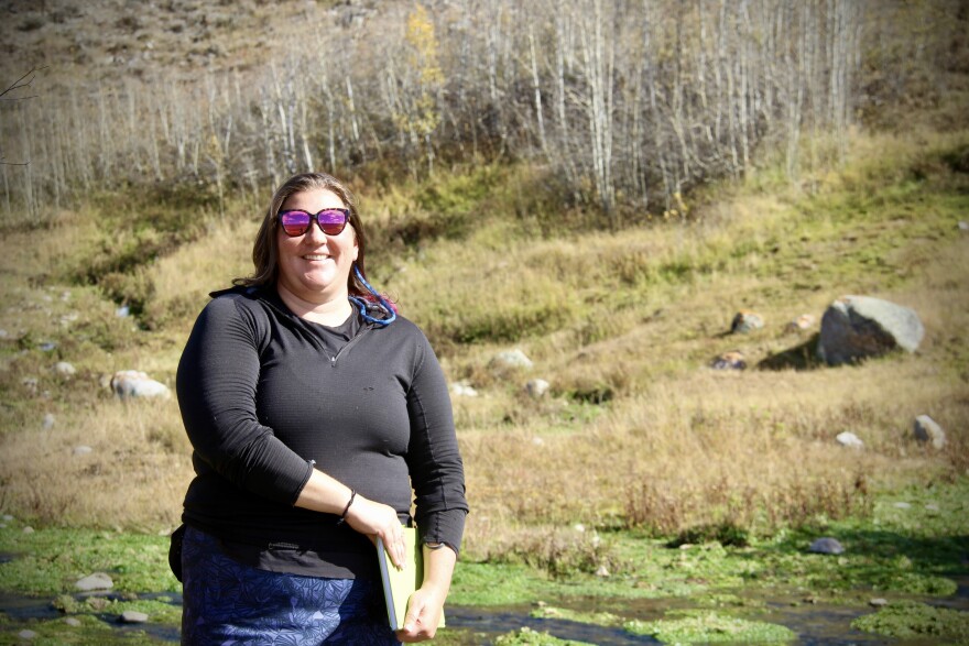 A woman faces the camera in front of a small body of water.