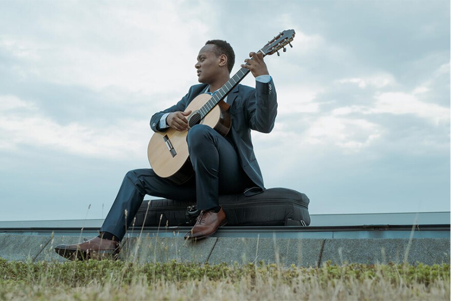 Raphaël Feuillâtre sits atop his guitar case holding his guitar, while looking into the distance