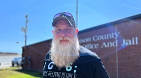 Dirk Harkins poses for a portrait. He has a long beard and wears a baseball hat and black t-shirt.
