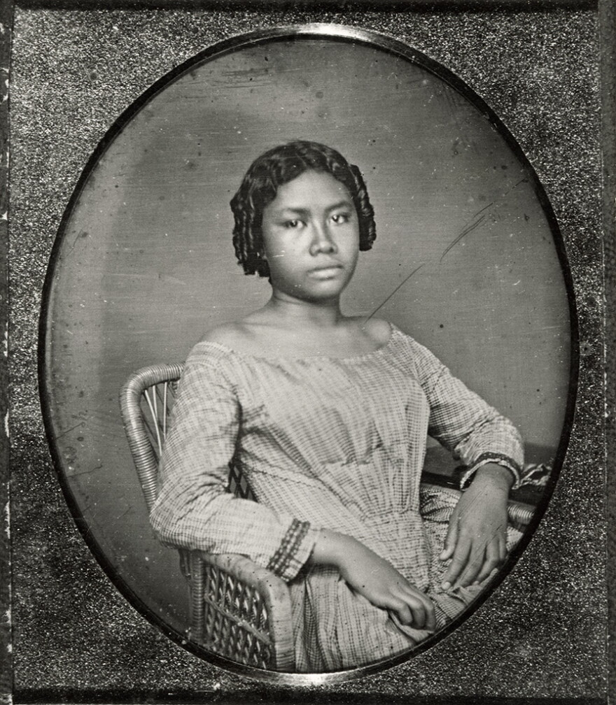 A black-and-white photo of Lili'uokalani, aged 15. Seh sits on a wicker chair with her hands in her lap.