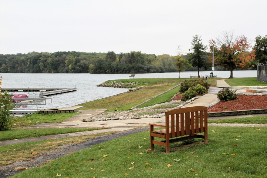 A wooden bench on a grassy lawn overlooking a lake with a boat dock and another bench on the distant shore.
