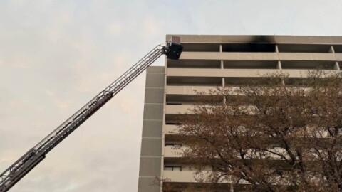 Fire trucks with extended ladder respond to a fire on the upper floor of a multi-story building, with black smoke visible from the windows. Trees and emergency lights are also visible in the scene.