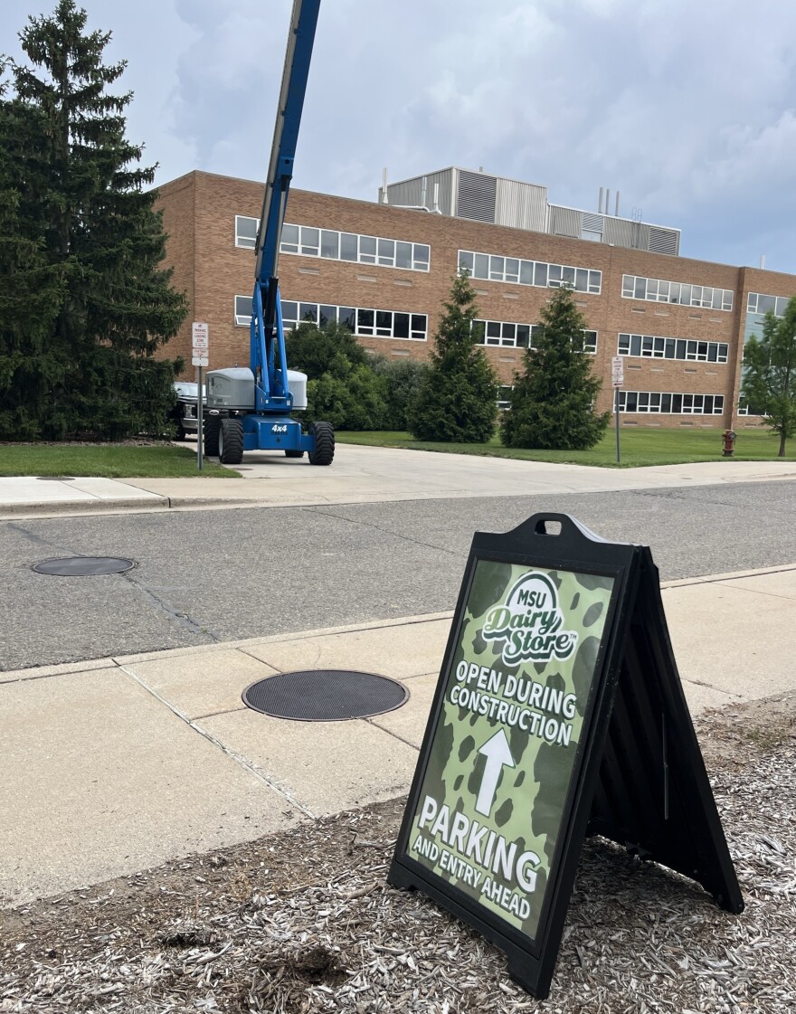 A sign on the sidewalk advertises the MSU Dairy Store, saying they're open during construction. An arrow directs to available parking.