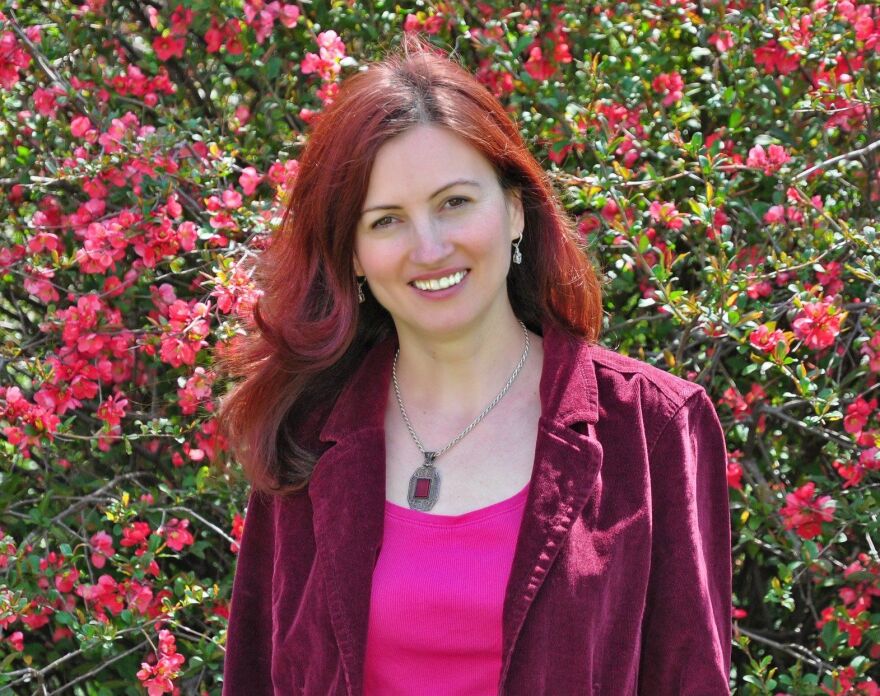 Lucia Cherciu wearing a red blazer and pendant necklace. Backdrop is a shrub with blooming flowers.
