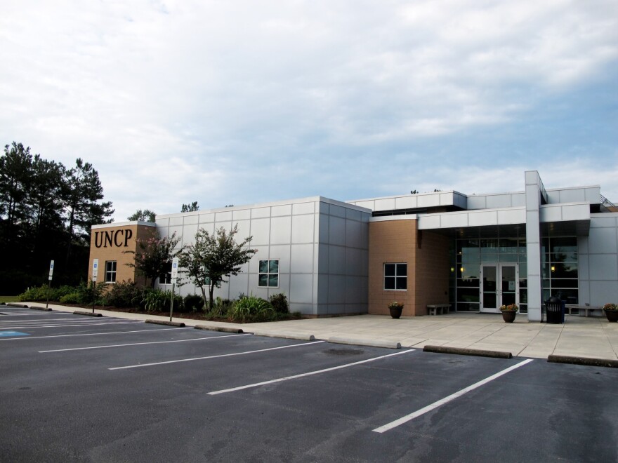 Photo showing a light gray and brown building. The letters UNCP are on the leftmost side of the building. 