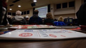 There are pamphlets on a table in the foreground and people sit in chairs watching a presentation.