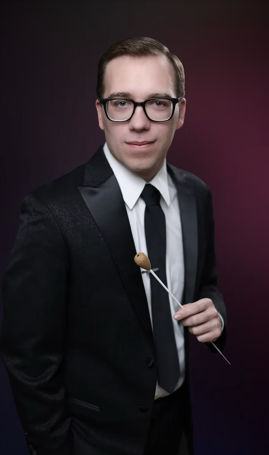 A young man with a conducting baton and glasses, dressed in formal black attire looks toward the camera