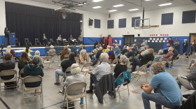 Attendees sit in brown folding chairs six feet apart inside the Mills Lawn elementary school gymnasium, to listen to the five challenger candidates for the Yellow Springs school board speak on a stage Tuesday night. The candidates are vying for three seats, and debated a school levy that would move the location of the school they were all sitting in.