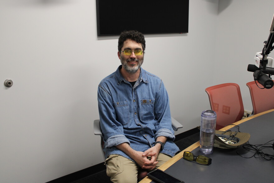 A man sits, smiling, in front of a studio microphone.