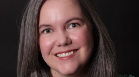 A rosy-cheeked woman with brownish-gray hair and a bright smile strikes a professional pose for the camera in a green and white dress. 