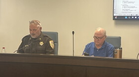An image of a man in sheriff's department uniform and another man sitting on a dais in a meeting of the McLean County Emergency Telephone System Board in McLean County.