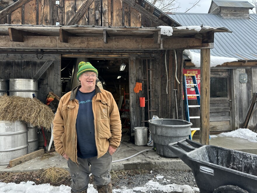 Man in green hat and work coat standing in front of his sugarhouse 