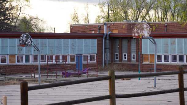 A dilapidated school building with boarded up windows surrounds a dirty paved playground, with dirty basketball hoops.