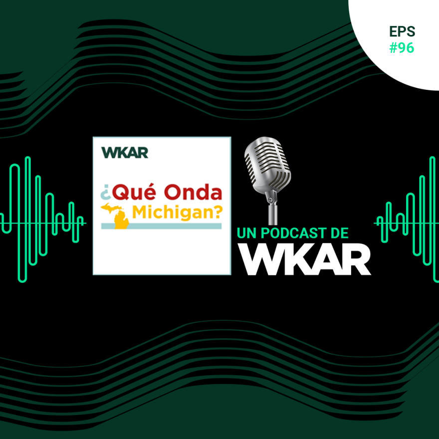 La imagen muestra un micrófono en un fondo negro. El micrófono es de color plateado y tiene una forma cilíndrica. Hay una rejilla en la parte delantera del micrófono para proteger la cápsula del micrófono. El micrófono está apoyado sobre un soporte de metal. En la parte superior del soporte hay una etiqueta que dice "EPS #96 WKAR ¿Qué Onda Michigan? UN PODCAST DE WKAR".
