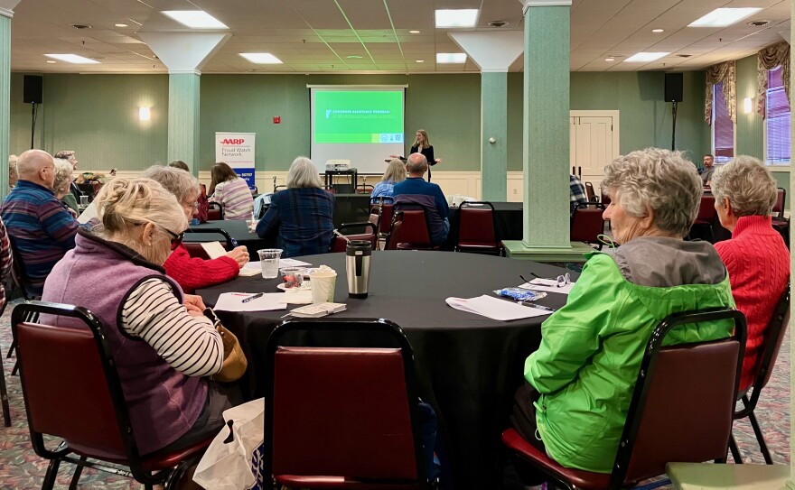 A group of older Vermonters listen to a speaker in a conference room.