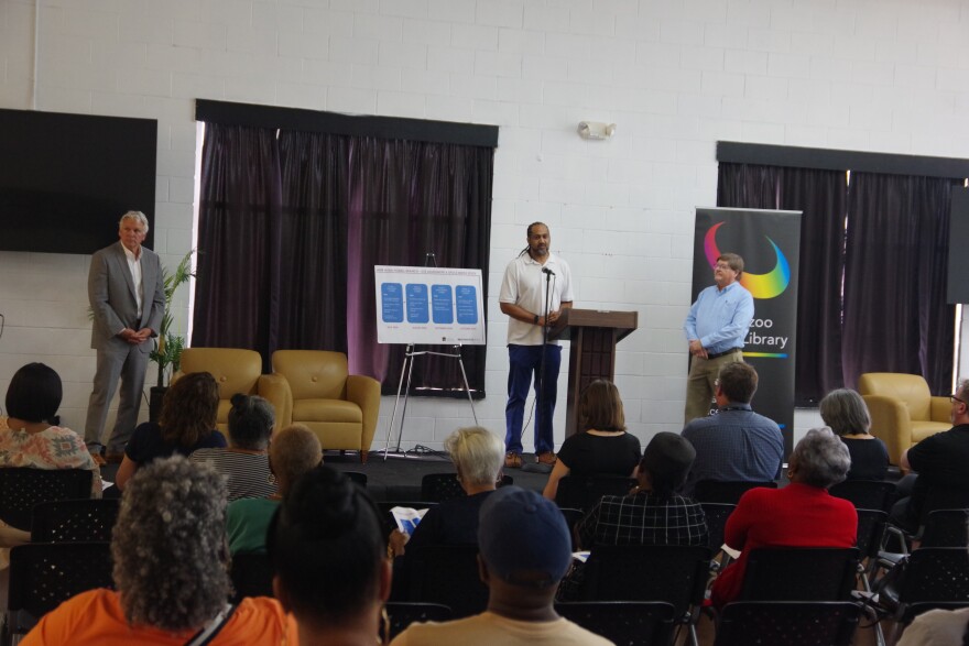 Three men stand on a stage, one at the podium in the middle, another to the far left of the image, and one to the right of the image. A poster sits on a stand to the right of the man at the podium, displaying information on the site assessment phases for a permanent Alma Powell location. Many community members sit in front of them. 