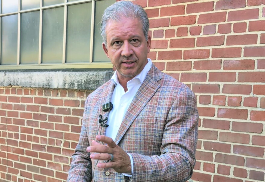 A man in a plaid suit stands in front of a brick wall and talks with reporters.