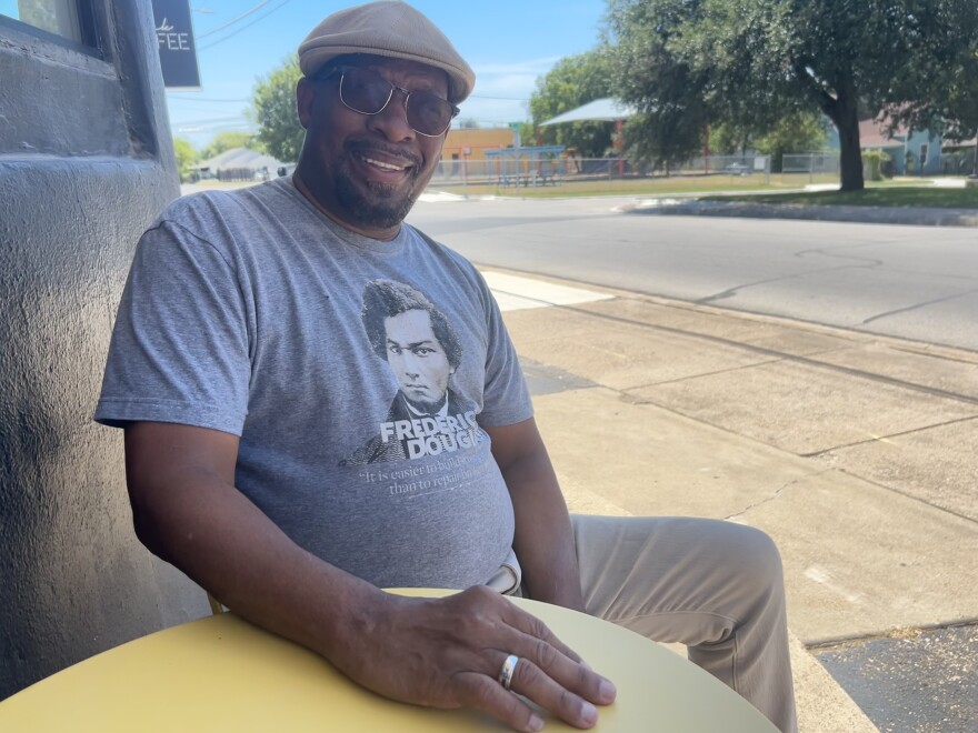 A photo of Wallace Bridges, a Black man wearing a cabbie hat and smiling for the camera while sitting at a small coffee shop table on a sidewalk. His t-shirt has a picture of Frederick Douglass.