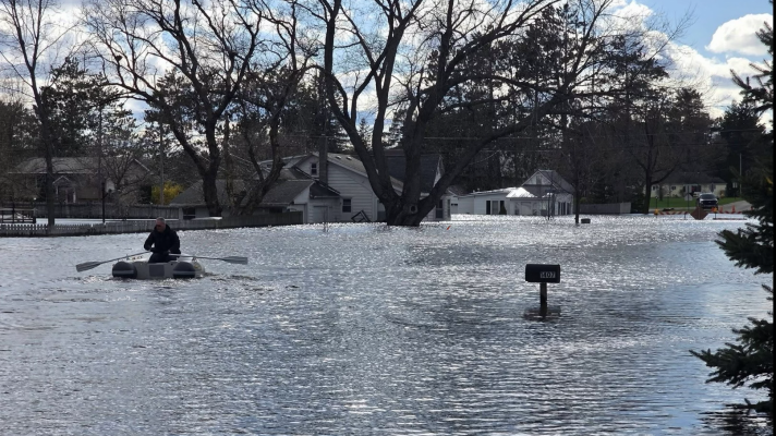Man paddles on a boat through flood waters in Cadillac, Michigan. Behind him are two white houses submerged in water. 
