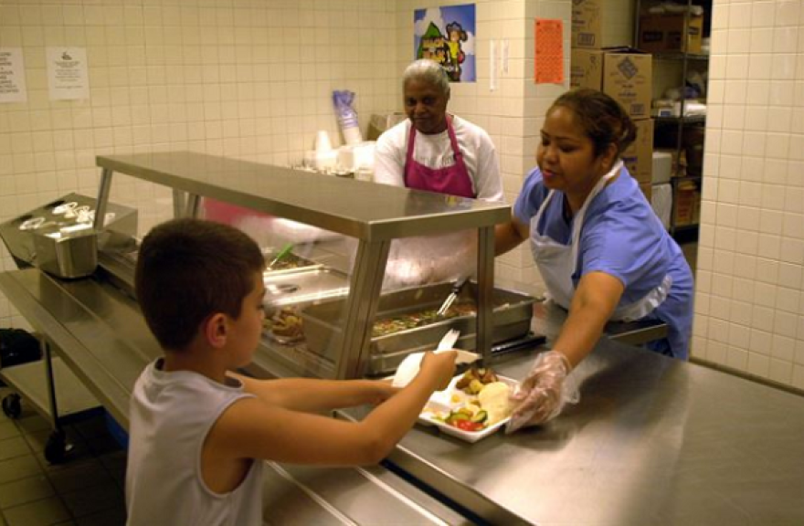 Two servers hand a tray of food to child in school cafeteria.