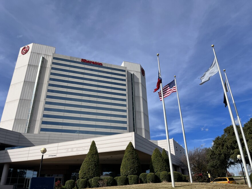 A 19-story hotel that has the red Sheraton "S" framed by leaf branches sits before a blue sky. There's a few flagpoles in frame, as well as an angular covered car area.