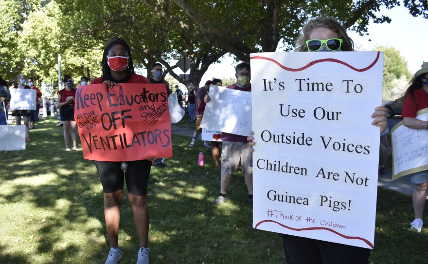 Two women hold signs that say, “It’s time to use our outside voices children are not guinea pigs!” and, “Keep our educators off ventilators.”