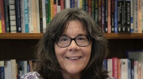 Ellen Birkett Morris standing in front of bookshelves. She has long dark hair with a bit of gray, and wears glasses.