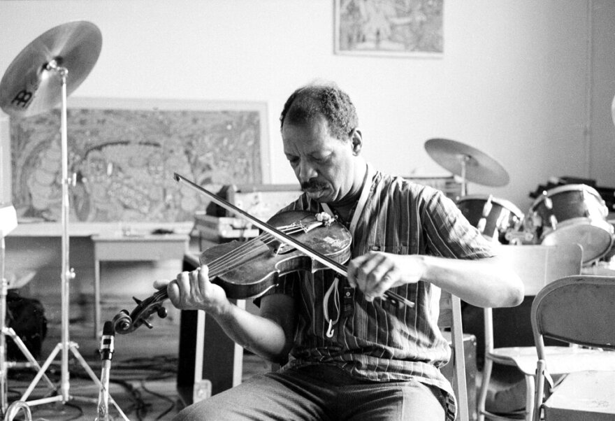 A black and white photo of a seated man playing a fiddle in a room filled with drums, cymbals and chairs.