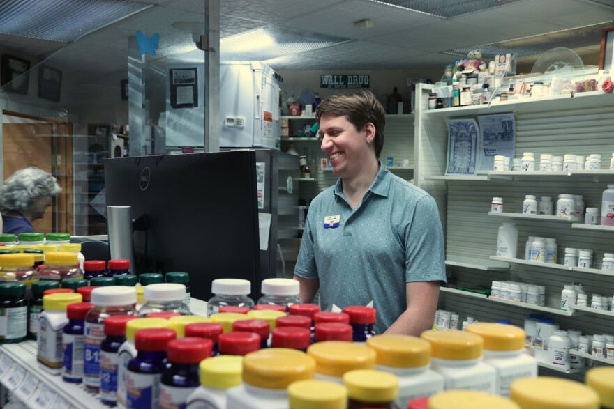 A man smiles and stands behind a counter filled with supplements and medications.