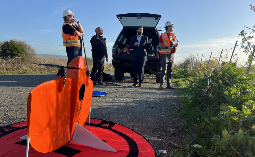 An orange fixed-wing drone in the foreground, four people in the background near a pickup truck. 