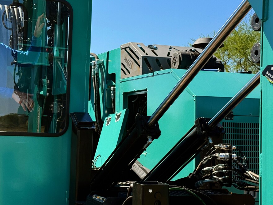 A close up of heavy machinery outside on a bright, sunny day. On the left, you can see a man in a blue shirt through the window of the cab. 