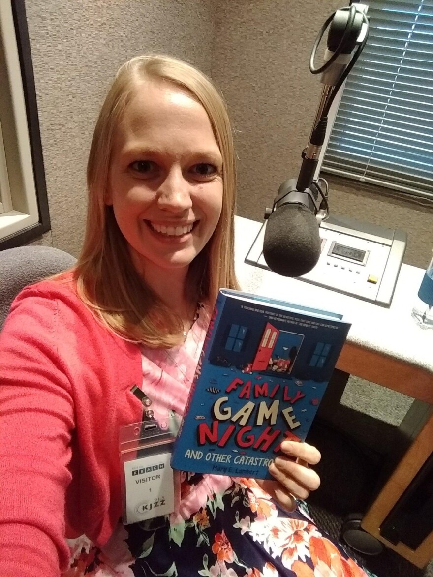 Author Mary Lambert at a radio recording studio in front of a microphone holding up her book Family Game Night and Other Catastrophes