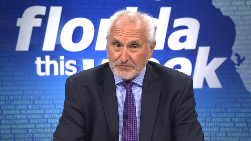 An elderly man with gray hair and a gray beard wearing a suit and tie with a blue background and white text saying "Florida This Week" in the background. 