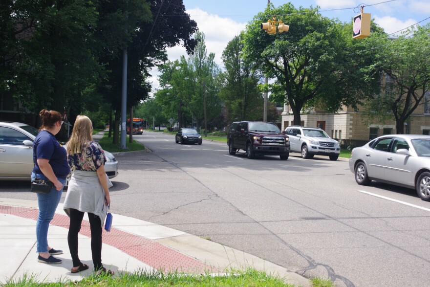 Two young women wait for several cars to clear an intersection on a wide one-way street.