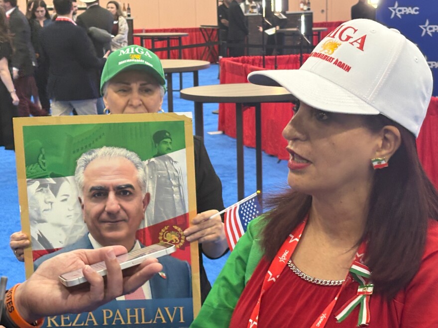 A woman wearing a "Make Iran Great Again" hat speaks with a reporter while a woman nearby can be seen holding  a poster of Reza Pahlavi at CPAC.