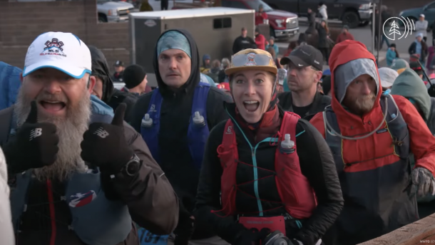 Runners wearing water bottle backpacks, hats, headbands, headphones, and long sleeves walk toward finish line and smile toward the camera.