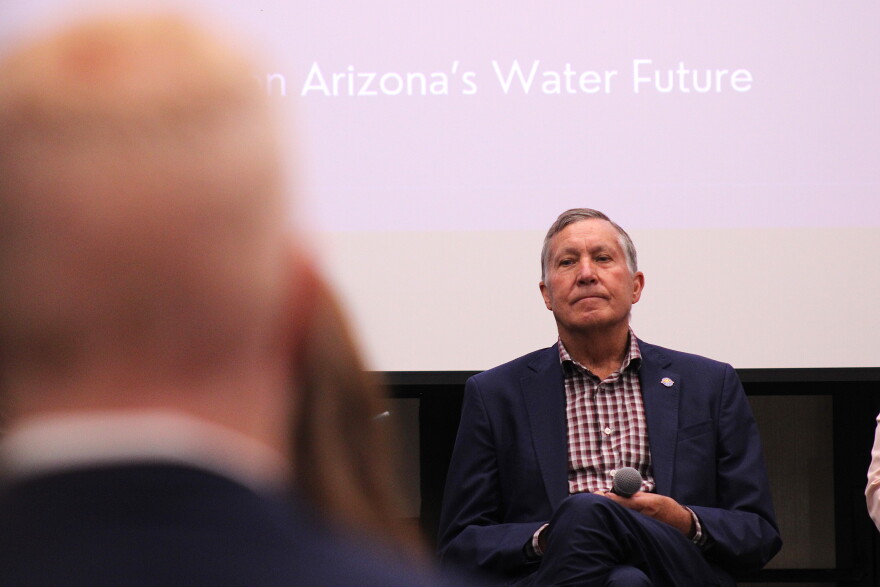 A man in a navy suit holds a microphone while looking out towards an audience from a stage. The words "Arizona's Water Future" appear on a screen behind him
