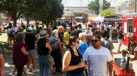 A large crowd of people line up to purchase tacos from a series of food trucks on a sunny summer day.