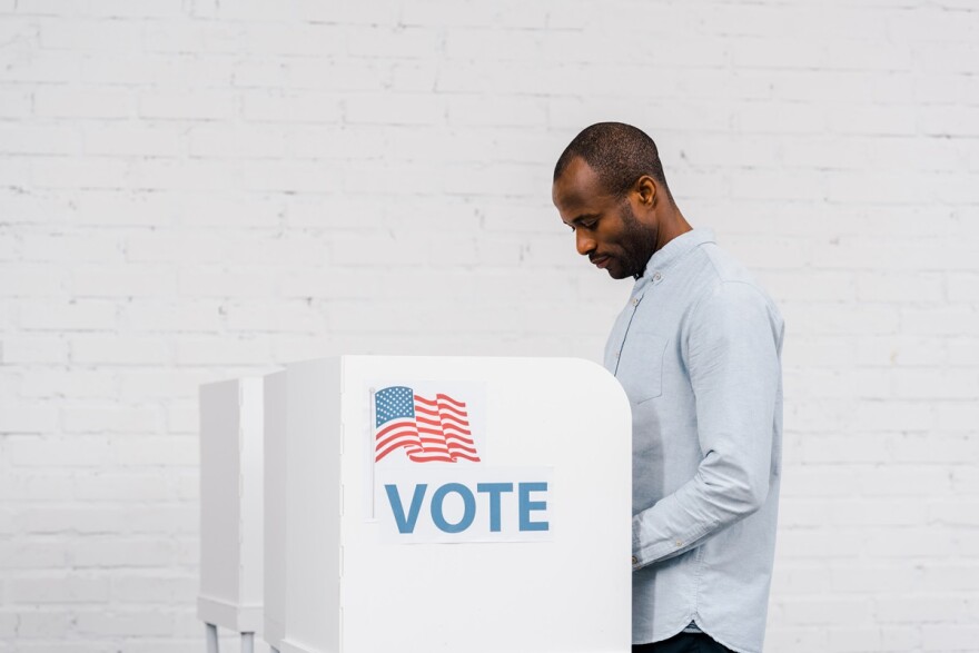 An African American mans votes at a booth with the word "vote" and an American flag on it