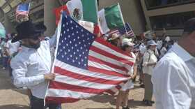 A man and a woman dressed in white hold an American flag as they march in a demonstration with other people. there are some Mexican flags visible behind them.