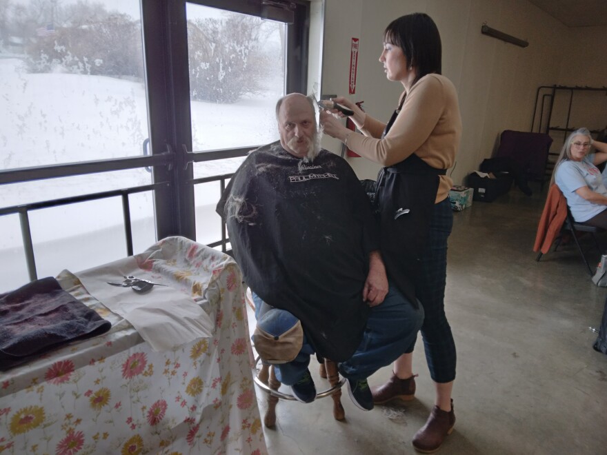 A man sits on a stool getting a hair and beard cut from a woman.