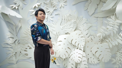 Lang Lang stands in a colorful shirt in front of a floral background, all painted white. He holds a yellow flower in his hand, and he smiles softly at the camera.