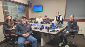 Six people gather around a table in a radio talk studio: a young woman front left has long brown hair and is wearing glasses, a blue fleece pullover and blue pants; a man front right has brown hair and is wearing a blue baseball cap, blue fleece pullover, jeans and grey shoes; a bald man back left has a grey mustache and is wearing glasses and a white polo shirt; a man back center is wearing a blue baseball cap and a blue fleece pullover, a man back right has short dark hair and is wearing glasses, a navy puffy vest, white hooded sweatshirt and blue jeans; a young woman at center wears her blonde hair pulled back and is wearing a blue fleece pullover, black pants and sneakers.
