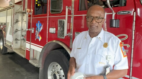 A uniformed fire department officer stands in front of a red fire truck holding a circular, white smoke alarm.