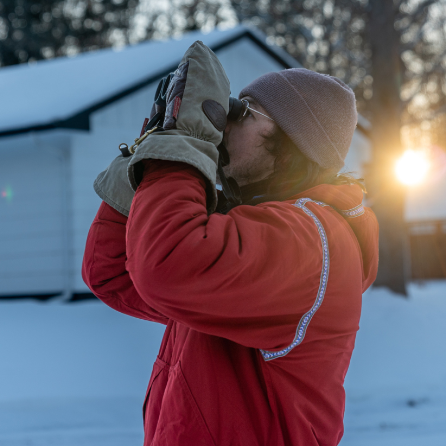 A man in a red coat and large mittens peers through binoculars on a winter morning.