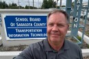 A gray haired man in gray collared shirt takes a selfie by a sign that reads "school board of Sarasota County."