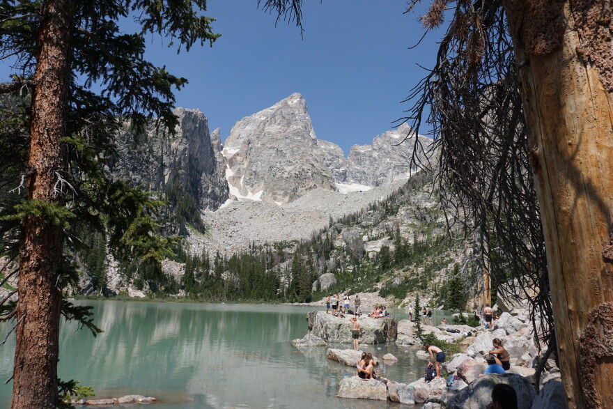 A rocky granite peak rises over an aquamarine lake. People sit and stand on rocks near the shore.