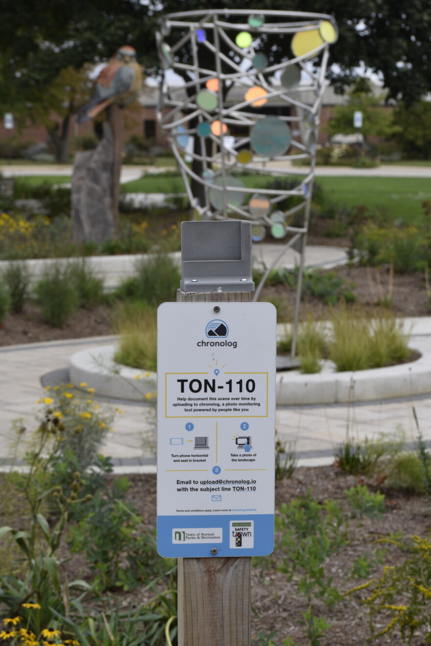 A Chronolog photo station labeled TON-110 stands in a garden with flowers, grasses, and a colorful metal sculpture in the background. A sitting statue of a person is visible further behind.